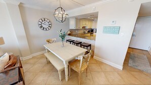 A dining area with a wooden table and bench, a clock, and a kitchen in the background. - Stunning BH 301A w/Seasonal Beach Service (Miramar Beach)