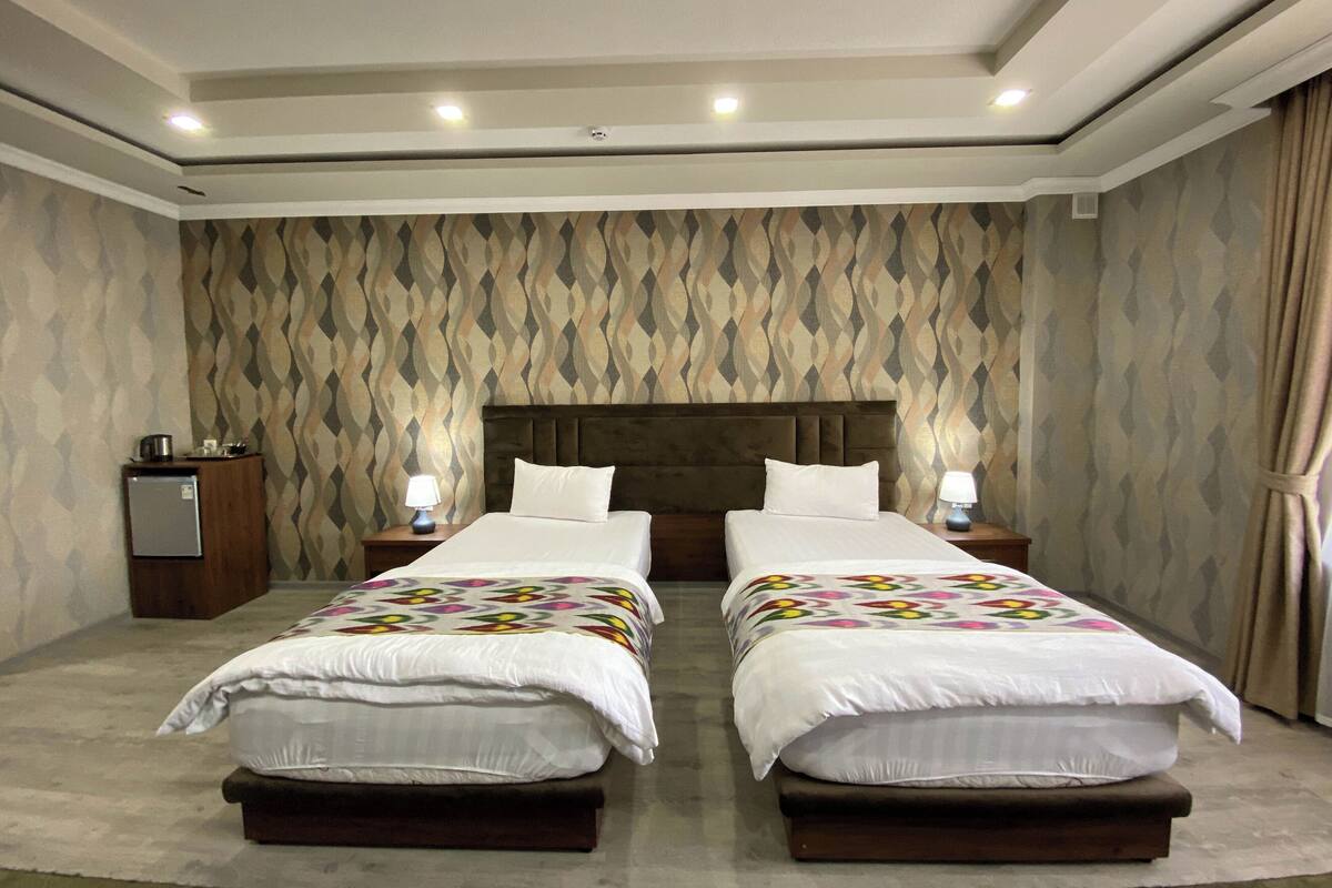 A double bed room with a patterned wallpaper, two bedside tables with lamps, and a minibar.