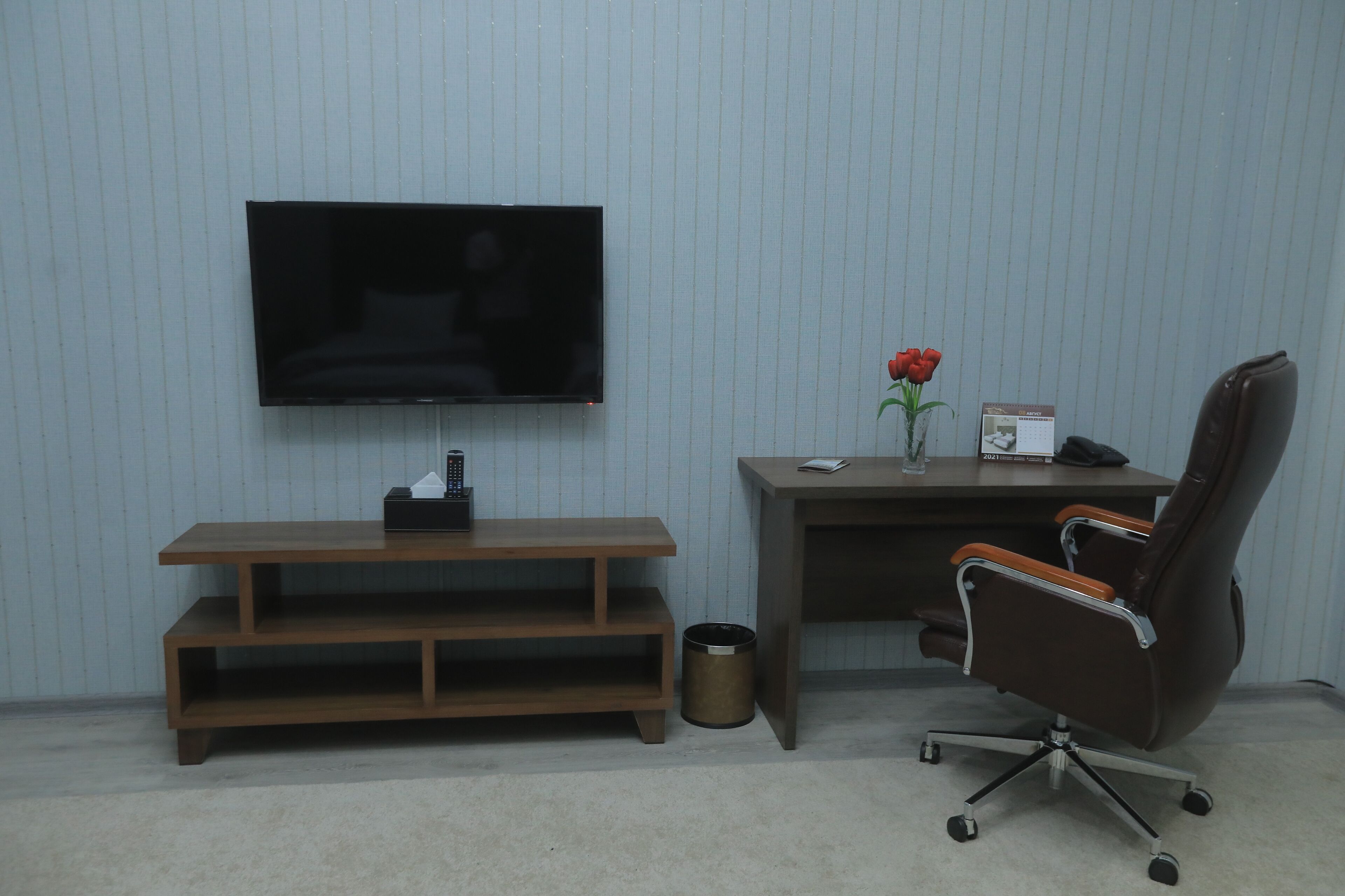 A room with a TV mounted on the wall, a wooden bench, a desk with a chair, a vase with a rose, and a trash can.