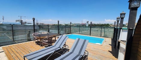 A rooftop pool area with lounge chairs and a table, surrounded by a fence and overlooking a cityscape.