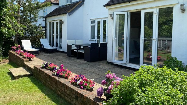 A two-story house with a patio area featuring outdoor furniture and a flower bed. - The Coniston Rooms (Kings Langley)