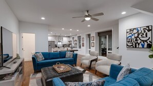 A modern living room with a blue sofa, a black ottoman, and a white armchair. There is a ceiling fan, recessed lighting, and a kitchen in the background.