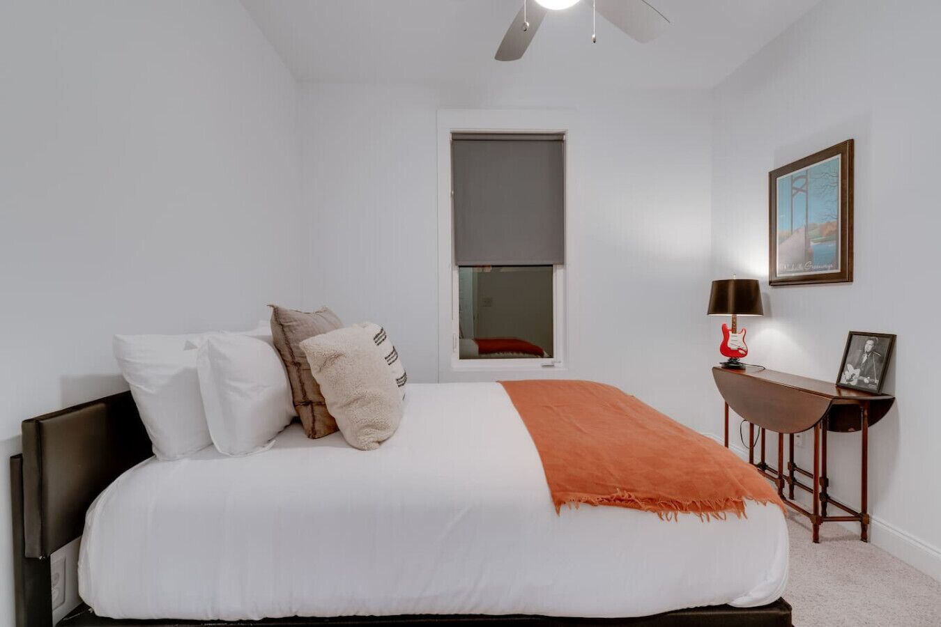 A bedroom with a large bed, a window, a desk with a lamp, and a framed picture on the wall.