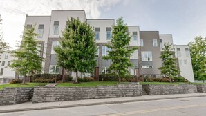 A modern apartment building with a uniform facade, multiple windows, and a landscaped front yard with trees and shrubs.