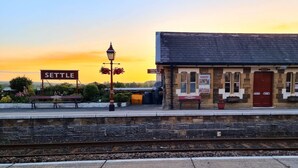 Una banchina di stazione ferroviaria con un cartello 'SETTLE', un cartello 'TRINDS TO CARLISLE' e un edificio con facciata in pietra e una porta rossa.