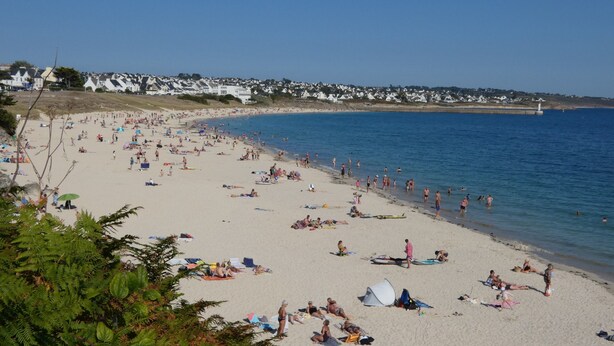 Une plage où des gens se prélassent au soleil et se baignent, un phare à l’horizon et des maisons le long de la côte.
