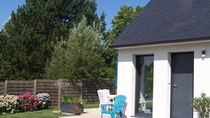 Une maison moderne avec un toit foncé, une façade blanche et une porte grise. On peut voir une chaise bleue à l’extérieur et un pot de fleurs près de la porte.