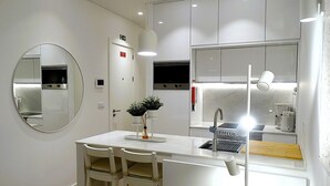 A modern kitchen with white cabinets, a round mirror, a small dining area with two chairs, and a coffee machine. - CA.Tanoeiros 1 (Funchal)