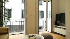 A modern living room with a sofa, a coffee table, a TV, and a balcony with a view of the building. - CA.Tanoeiros 1 (Funchal)