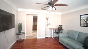 A living room with a wooden floor, a ceiling fan, a TV mounted on the wall, a green sofa, and a dining area with a table and chairs. - Federal Ct- Charming 3-bedroom near Ag Center and MTSU in Murfreesboro (Murfreesboro)