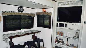 Una habitación pequeña con un escritorio, dos taburetes negros, un televisor y una estantería con varios objetos.