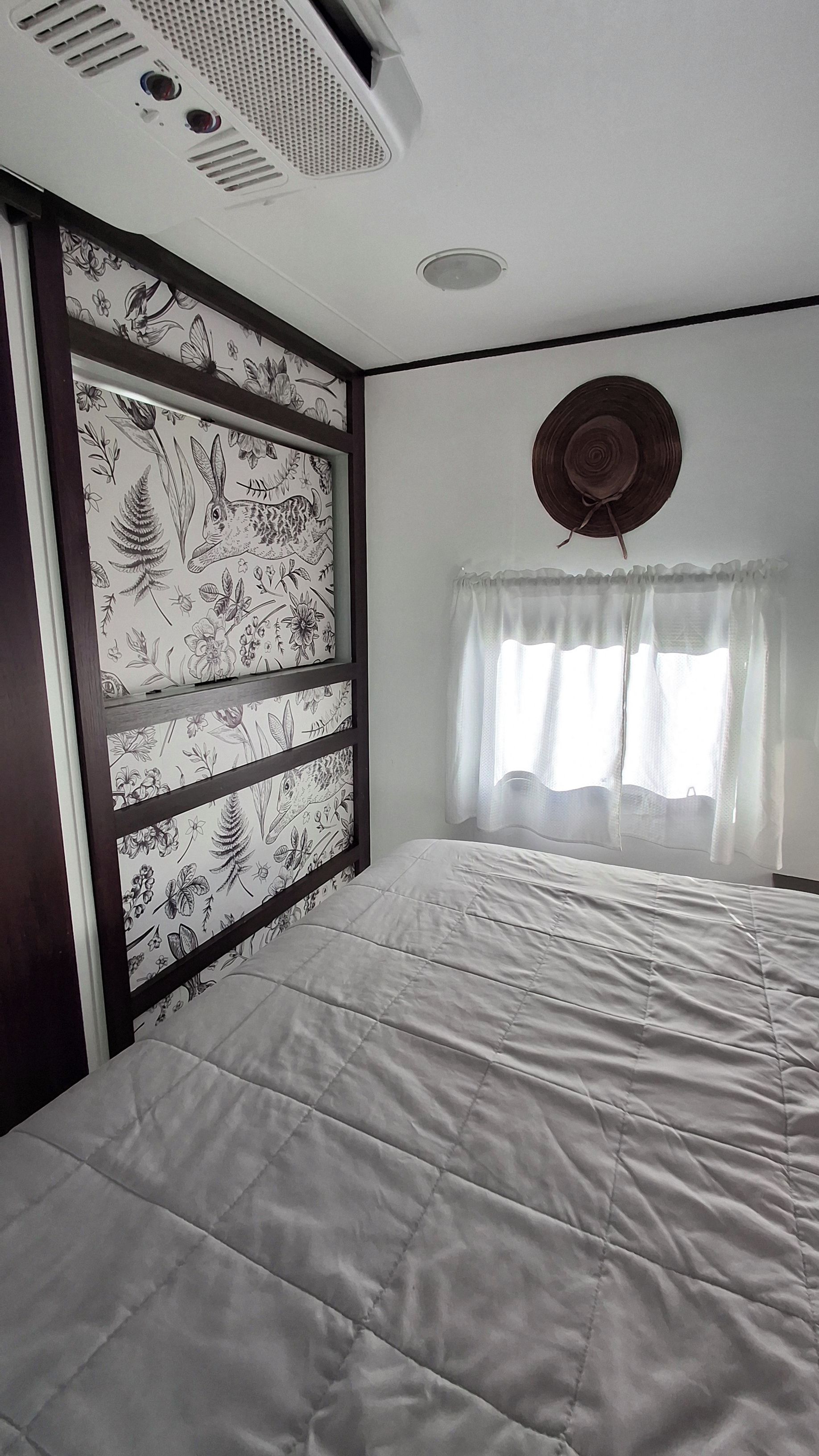 Ein Schlafzimmer mit einem Bett, einem Fenster mit Vorhängen, einer Wand mit Blumenmuster und einem Deckenventil.