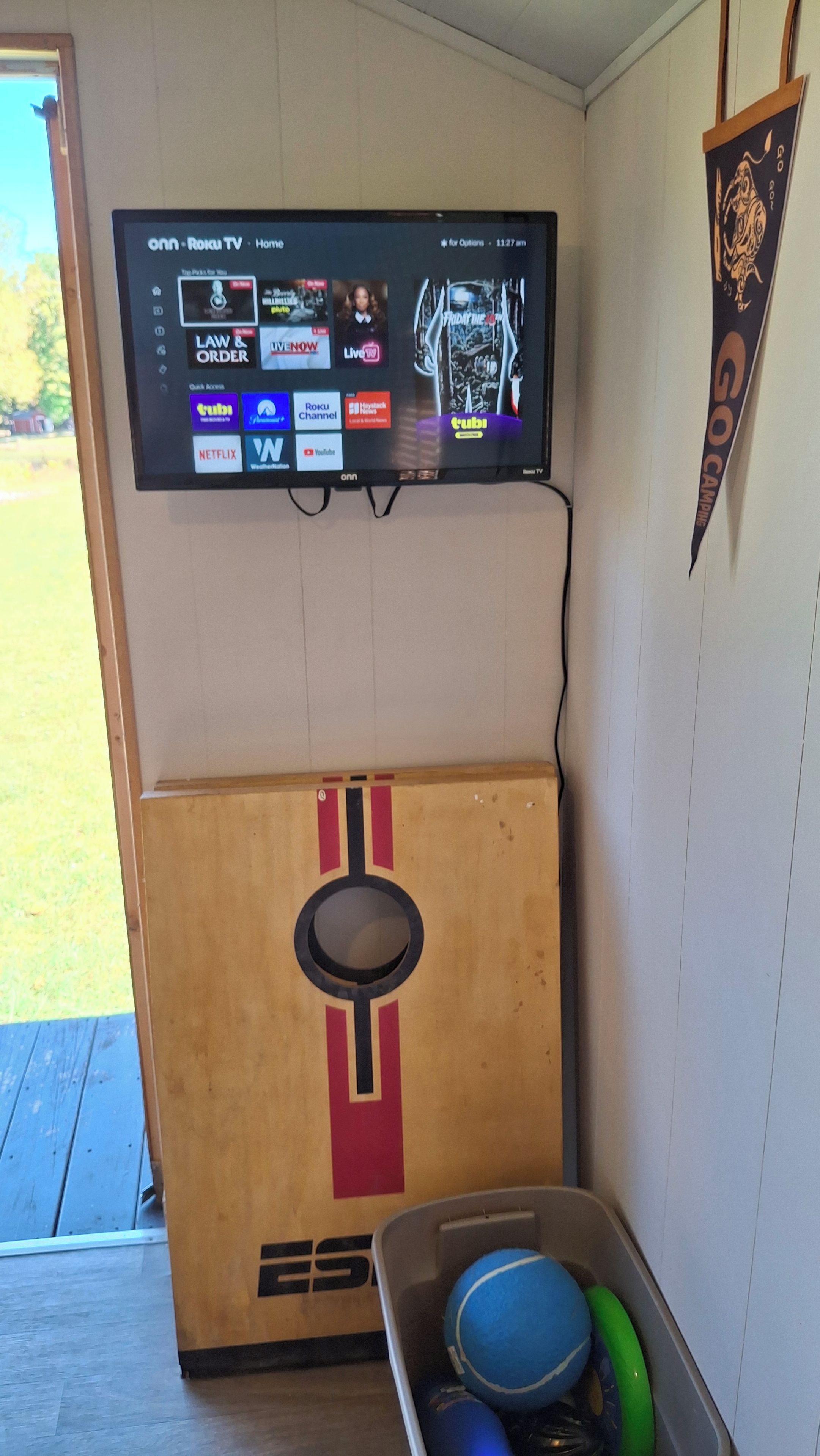 A television mounted on a wall displaying various streaming services. Below the TV, there is a wooden cabinet with a circular cutout and a metal container holding sports balls.