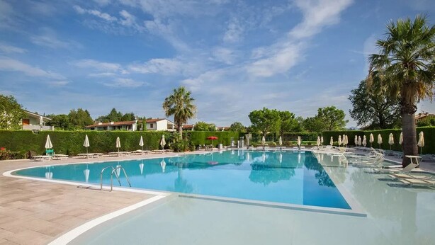 A large, clear swimming pool with lounge chairs and umbrellas, surrounded by greenery and buildings. - Apart Dina Lazise, pool, parking, WIFI, KLima, garden, laundry package (Colà)