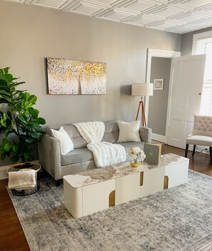 Un salon avec un canapé gris, une table basse blanche, une grande plante et un tableau accroché au mur.