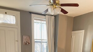 Un dormitorio con cama, mesita de noche, cómoda, ventana con cortinas, ventilador de techo y puerta.