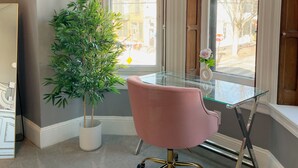 Una oficina en esquina con escritorio de vidrio, una silla rosa, una planta en maceta y ventanas grandes con contraventanas de madera.