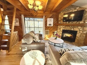 A cozy cabin interior with a stone fireplace, wooden beams, a comfortable sofa, and a television mounted on the wall.