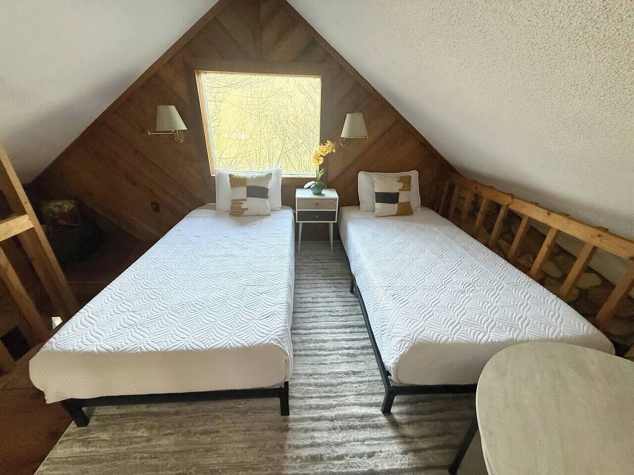 A small, attic-style bedroom with two single beds, a nightstand, a lamp, and a window with a view.