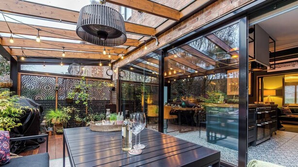 Une terrasse extérieure moderne, avec une table en bois dressée pour les repas, entourée de plantes en pot et protégée par une paroi en verre.