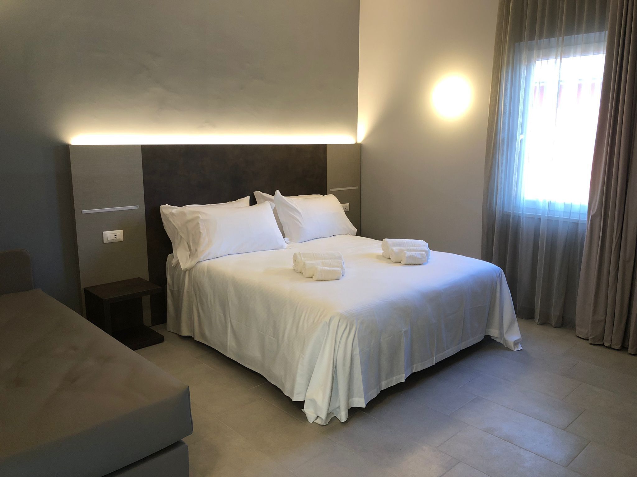 Una habitación de hotel moderna con una cama bien hecha, una mesita de noche y un ventanal con cortinas.