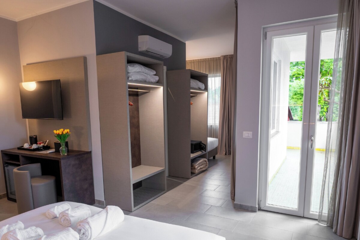 A hotel room with a bed, a desk with a flat-screen TV, a wardrobe, and a sliding glass door to the outside.