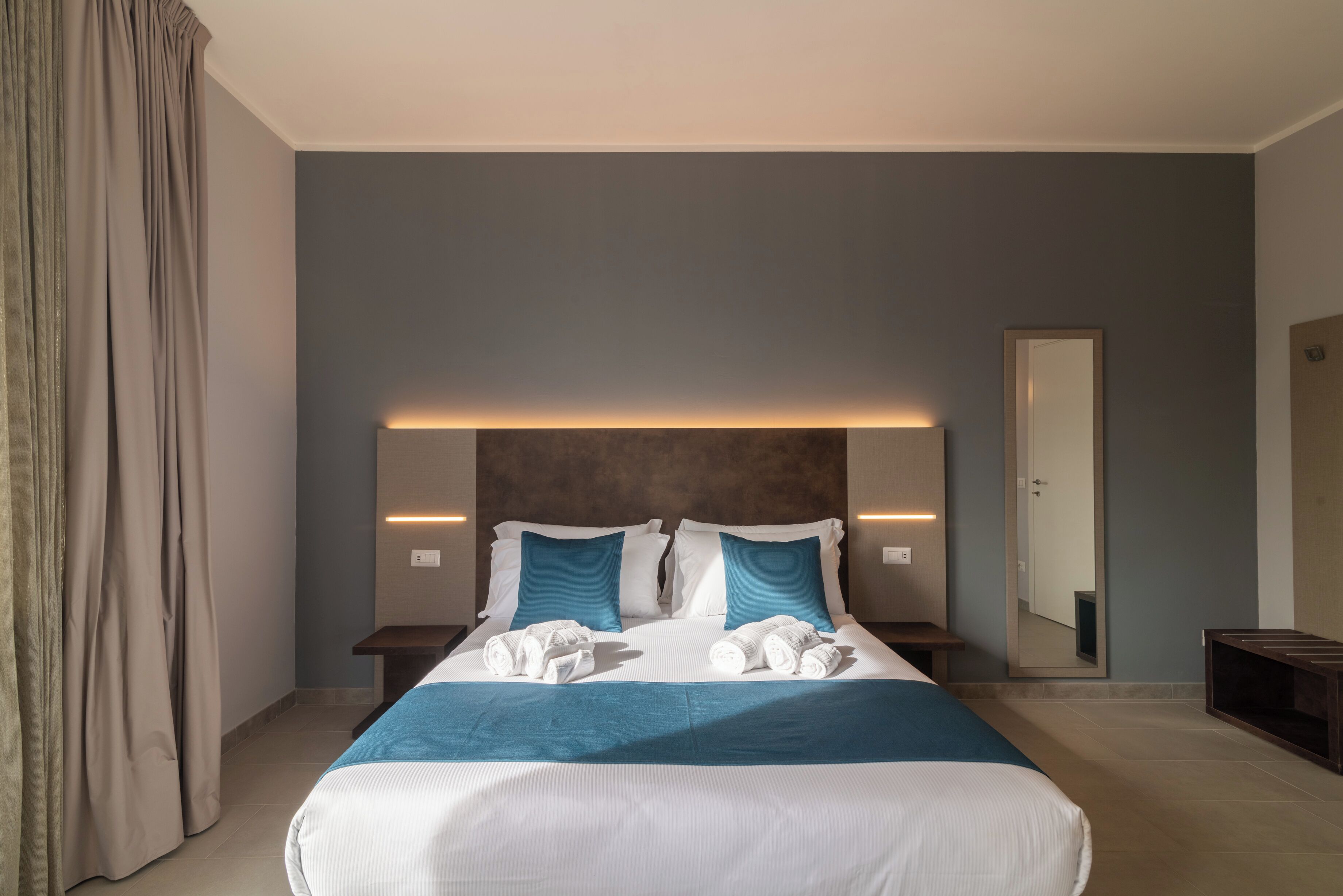 A modern hotel room with a large bed, two bedside tables, a nightstand, a mirror, and a curtain.