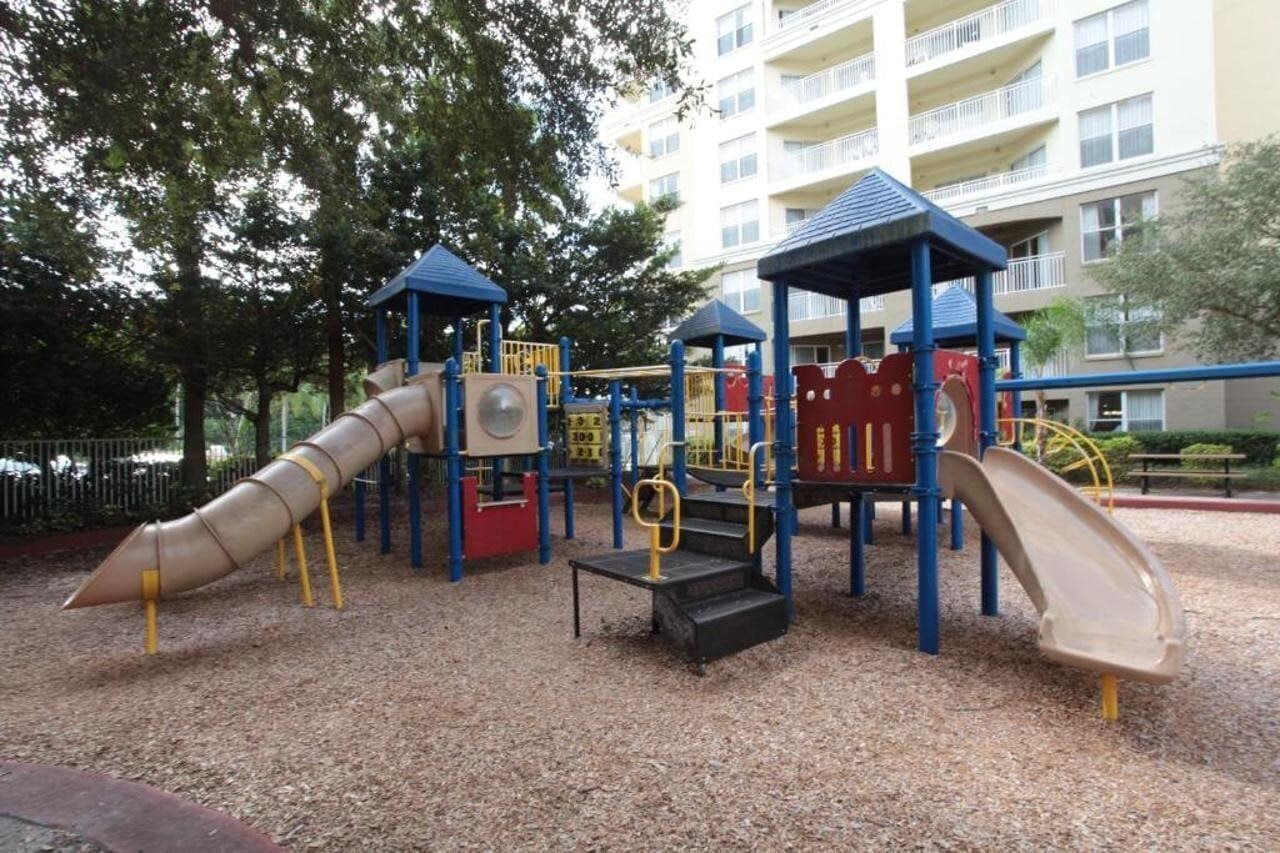 A playground with slides and climbing structures, surrounded by trees and a building in the background.