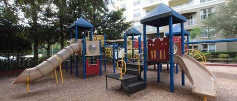 A playground with slides and climbing structures, surrounded by trees and a building in the background.