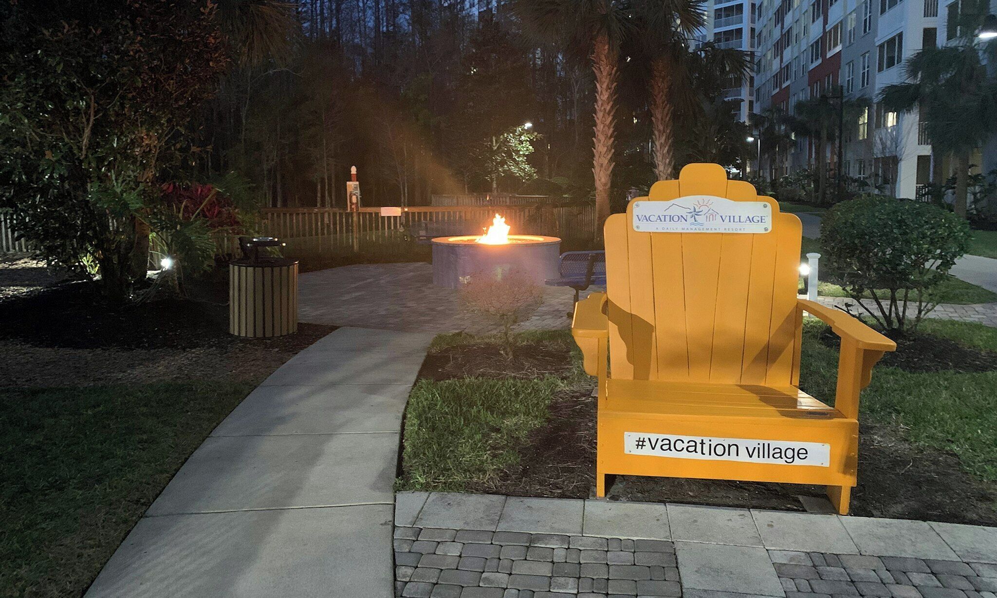 Ein Weg führt zu einer Feuerstelle und einem Holzstuhl mit der Aufschrift 'Vacation Village'.
