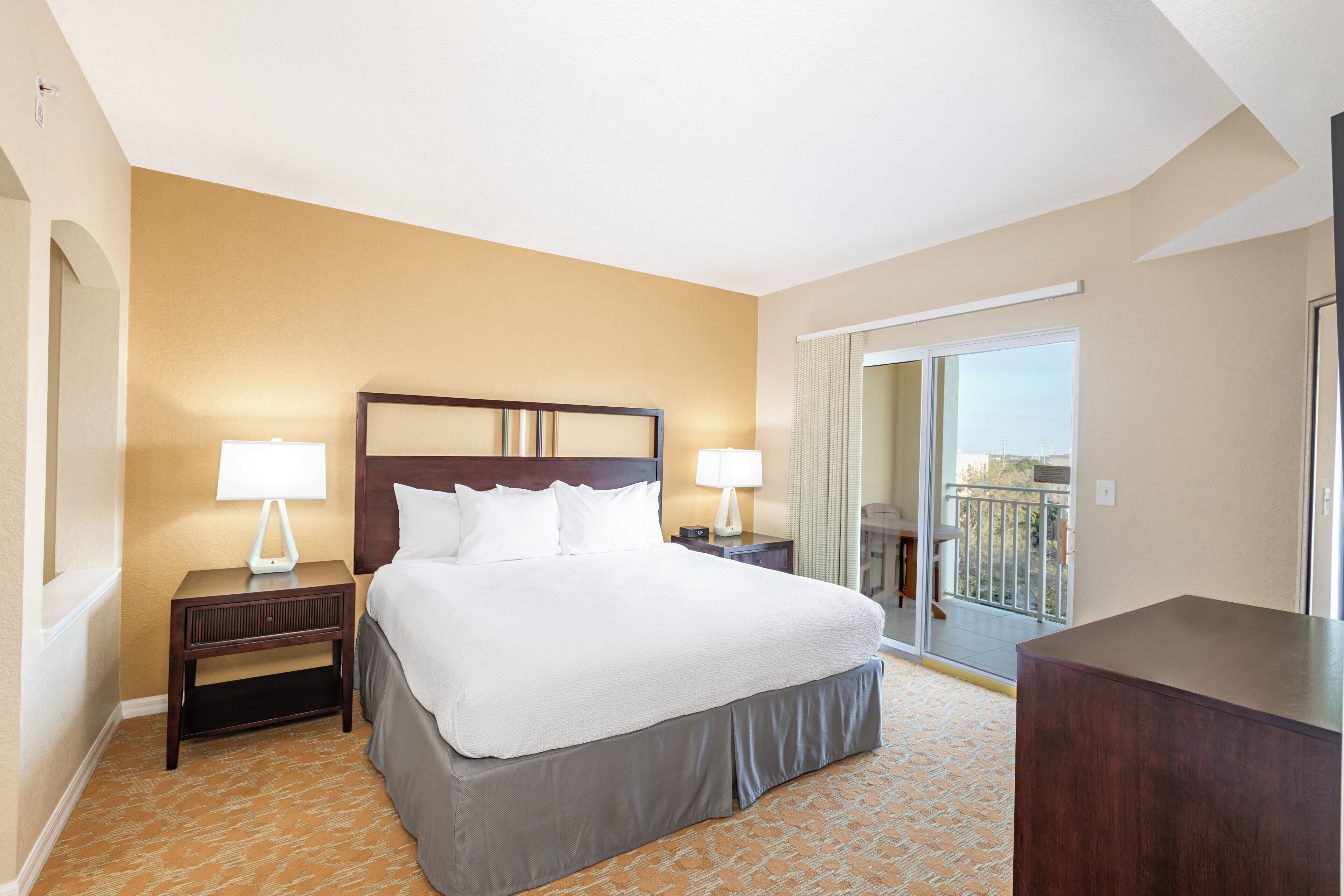 A hotel room with a large bed, two bedside tables with lamps, a view of a balcony, and a door leading to an outdoor area.