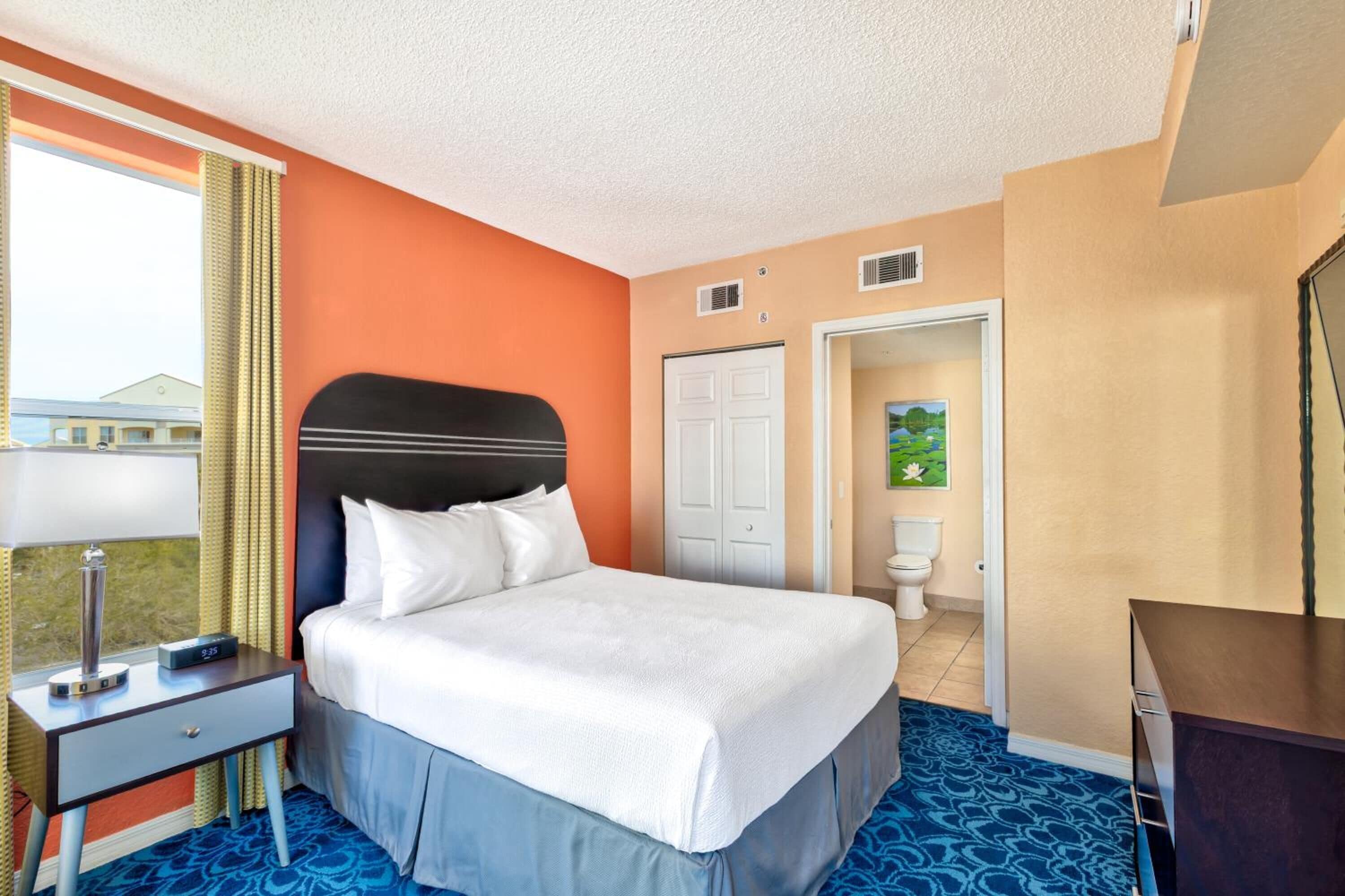 Une chambre d’hôtel avec un lit, une table de chevet, un bureau, une fenêtre avec des rideaux, une porte et un tableau au mur.
