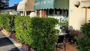 A hotel named HOTEL ETRUSCO with a green awning, outdoor seating area with tables and chairs, and potted plants lining the walkway.
