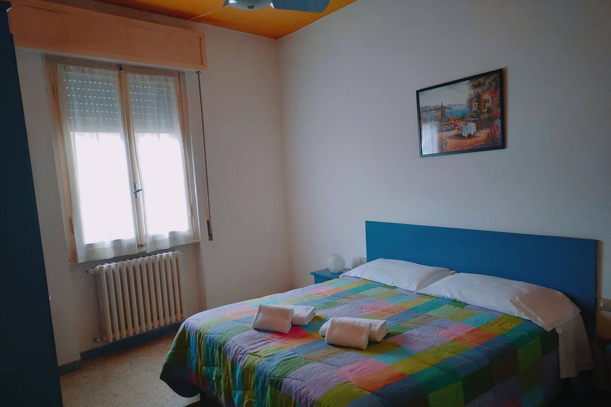 Ein Schlafzimmer mit Bett, Fenster mit Jalousien, Heizkörper, Nachttisch und einem Bild an der Wand.