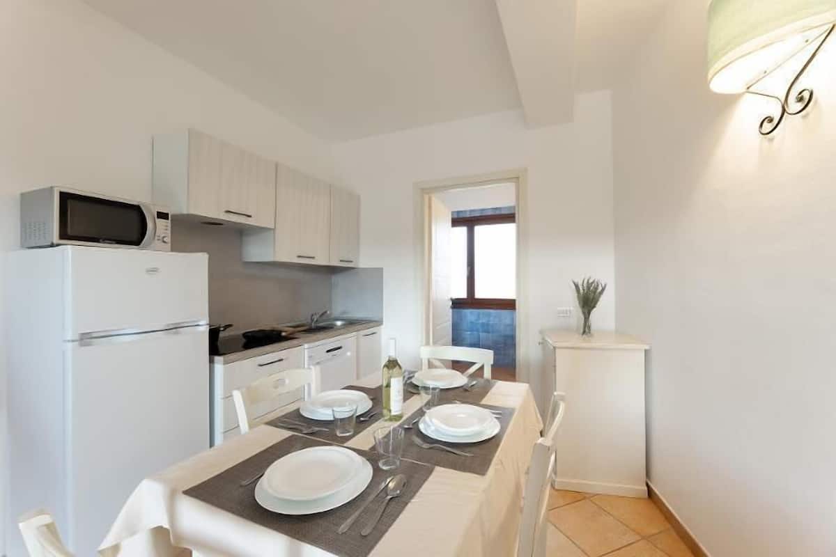Cocina compacta con armarios blancos, microondas y una mesa de comedor preparada para dos personas, con platos, cubiertos y una botella.