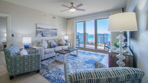 A spacious living room with a large window offering a view of the ocean and high-rise buildings. There is a ceiling fan, a sofa, a coffee table, and patterned chairs.