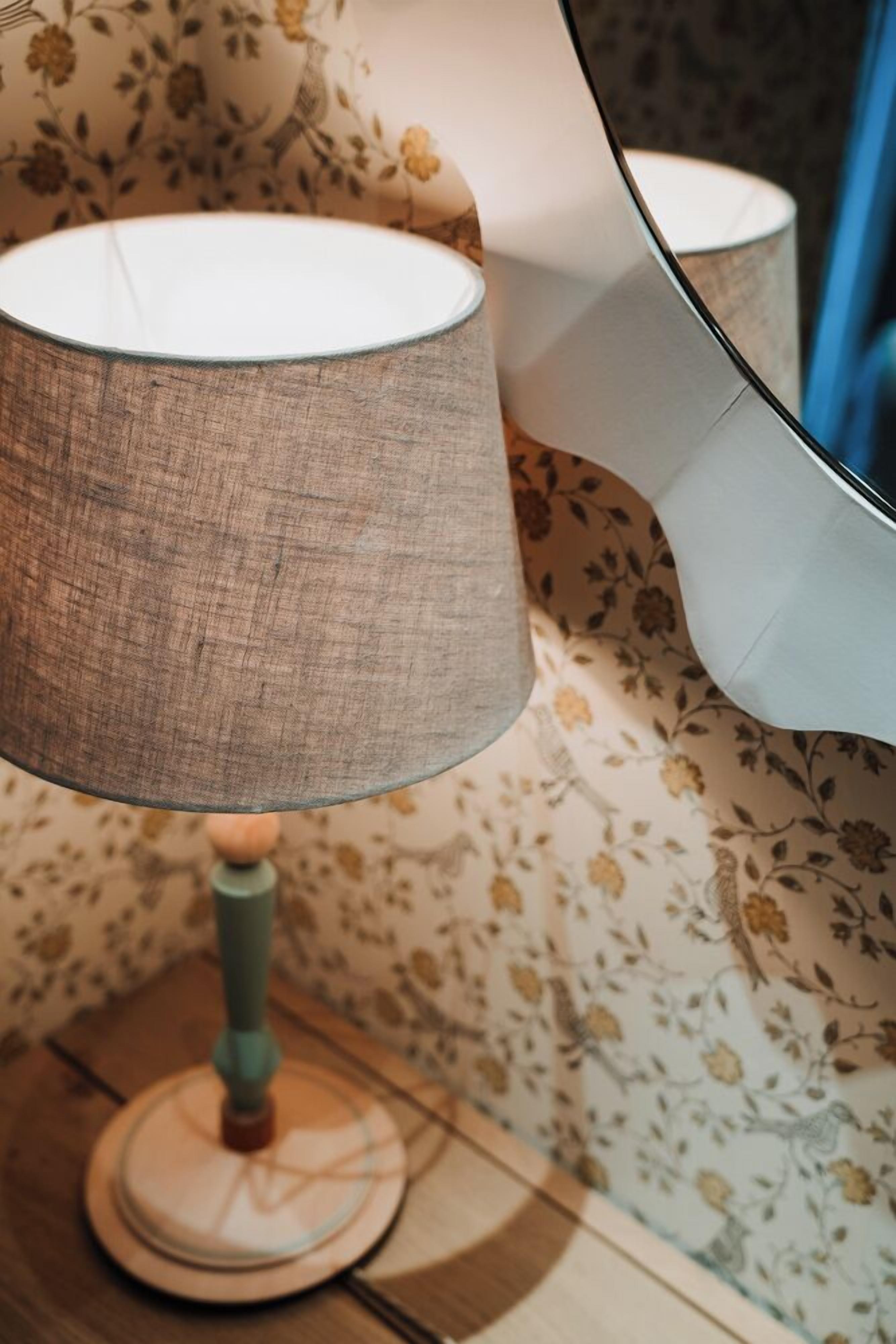 A lamp with a textured shade on a wooden table against a floral wallpaper background.