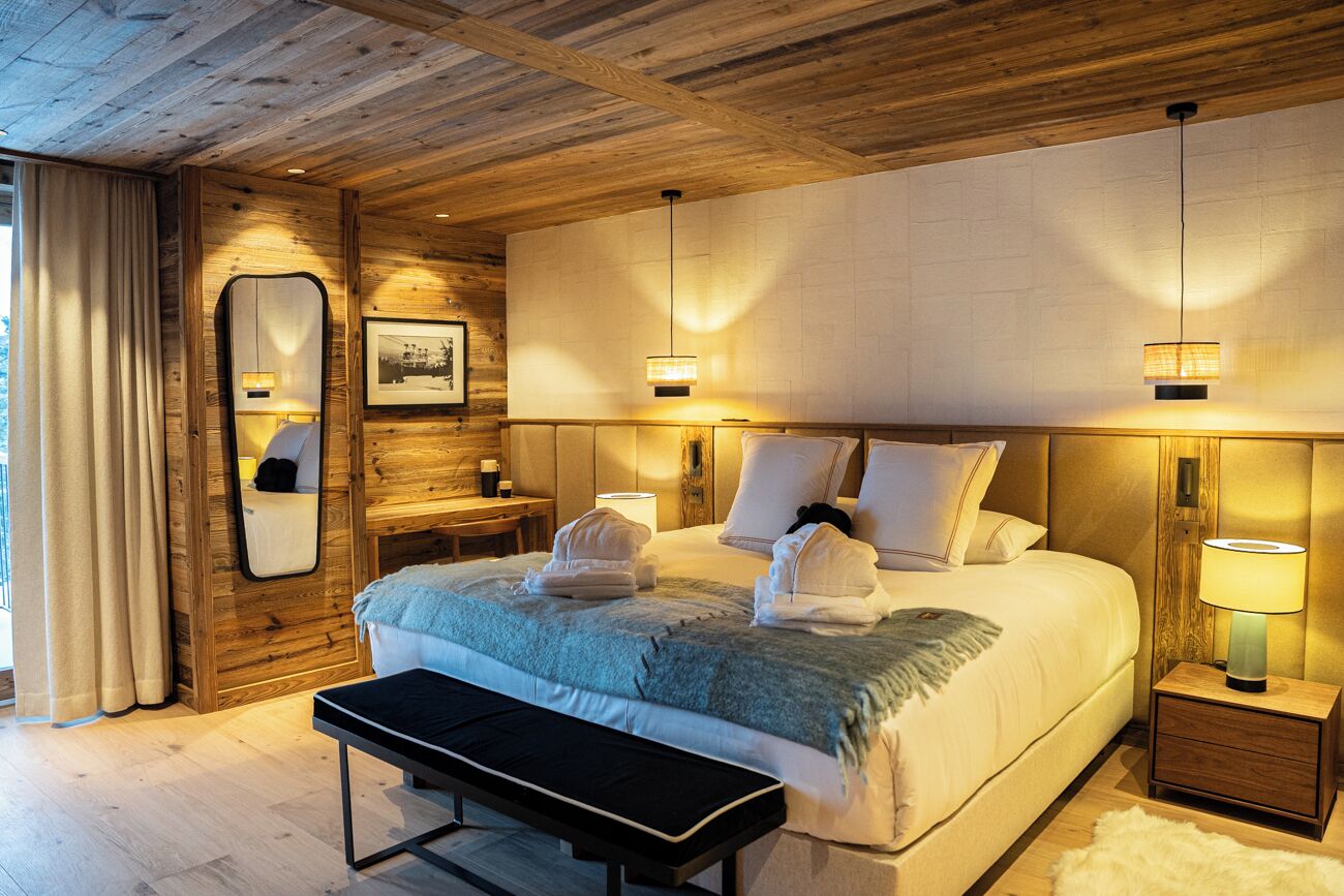 Een moderne slaapkamer met een houten plafond, een groot bed met kussens en dekbed, nachtkastjes met lampen, een bankje en een spiegel aan de muur.