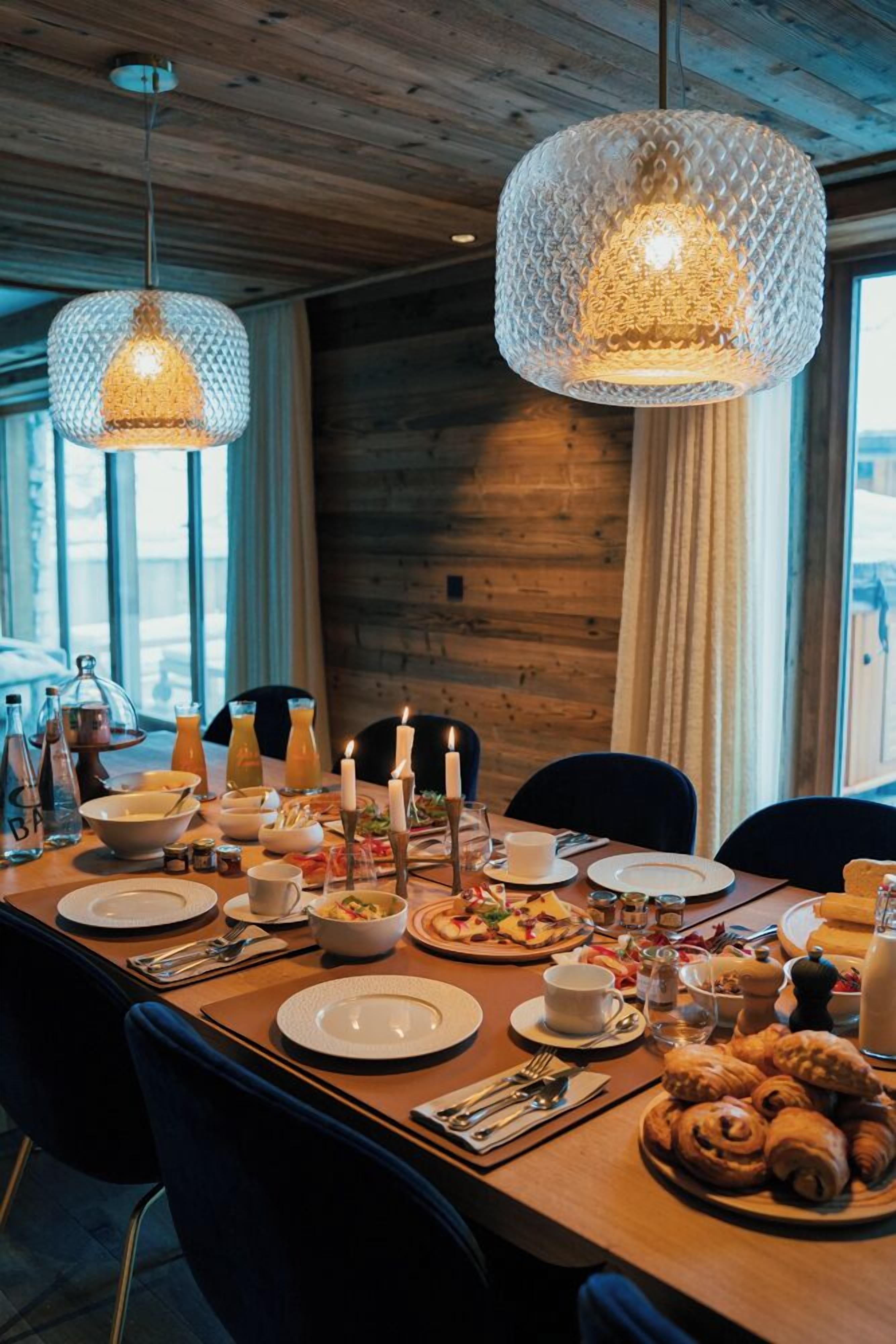 Een eettafel gedekt met borden, bestek, kaarsen en gebak, met hangende kristallen kroonluchters en houten wanden.