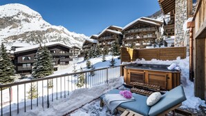 A snow-covered balcony with a hot tub, lounge chairs, and a view of mountain cabins. - Silverstone by Alpine Resorts (Val-d'Isere)
