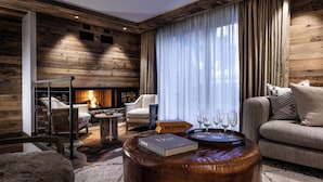 A cozy living room with a fireplace, leather ottoman, and a bookshelf with books and glasses. - Silverstone by Alpine Resorts (Val-d'Isere)