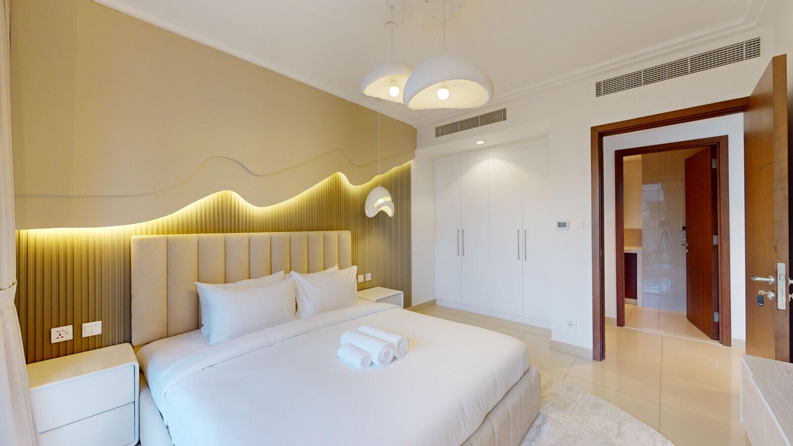 A modern bedroom with a large bed, bedside tables, a wardrobe, and a door leading to another room.