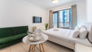 Une chambre d’hôtel moderne avec un canapé vert, un lit blanc, une table à manger avec des assiettes et des verres, un téléviseur fixé au mur et un balcon avec des rideaux.