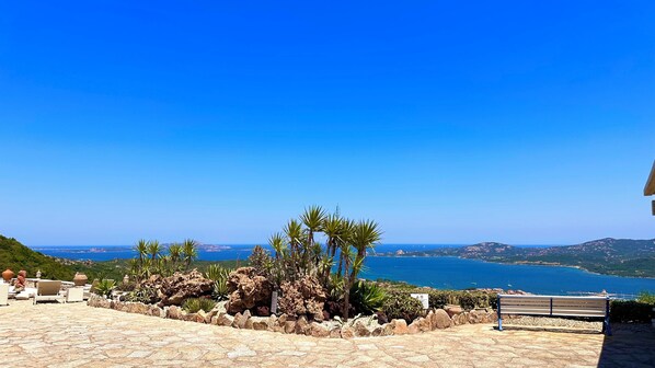 A patio with stone flooring, a bench, and a view of the sea and distant hills.