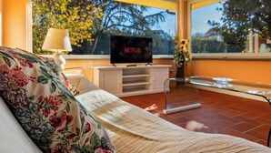 A living room with a sofa, a TV on a stand, a glass table, and a window with a view of trees. - *Elegant Villa*with Private Park-10 min from LUCCA (Capannori)