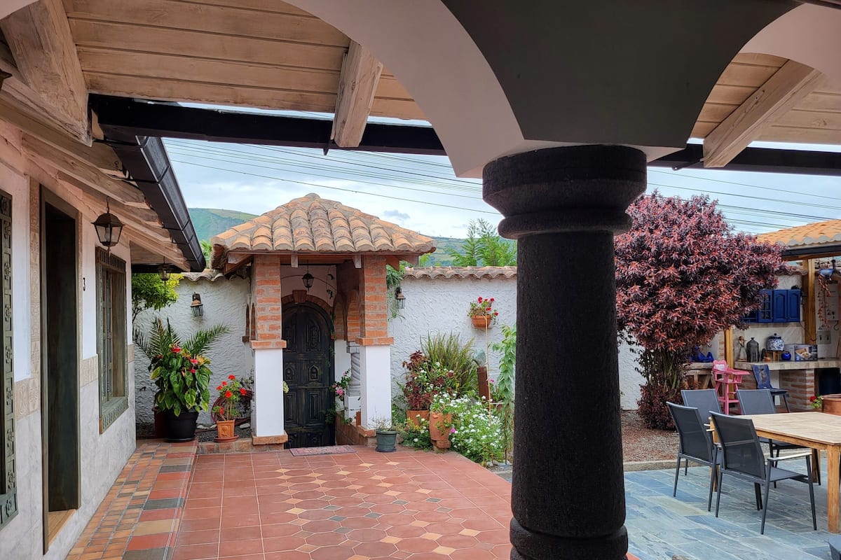 A courtyard with a tiled floor, a wooden pergola, a stone column, and a dining area with chairs and a table.