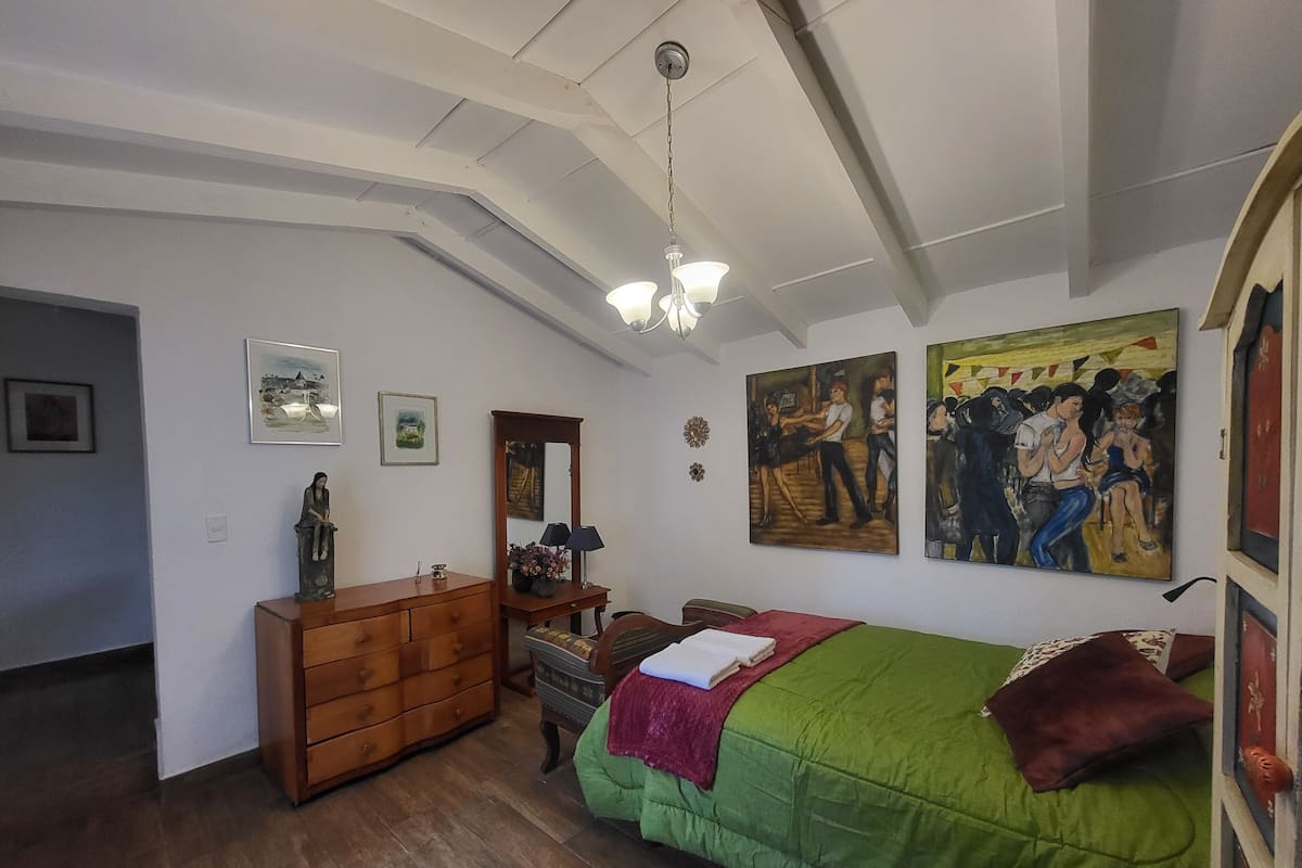 A bedroom with a green bed, wooden dresser, and two paintings on the wall.