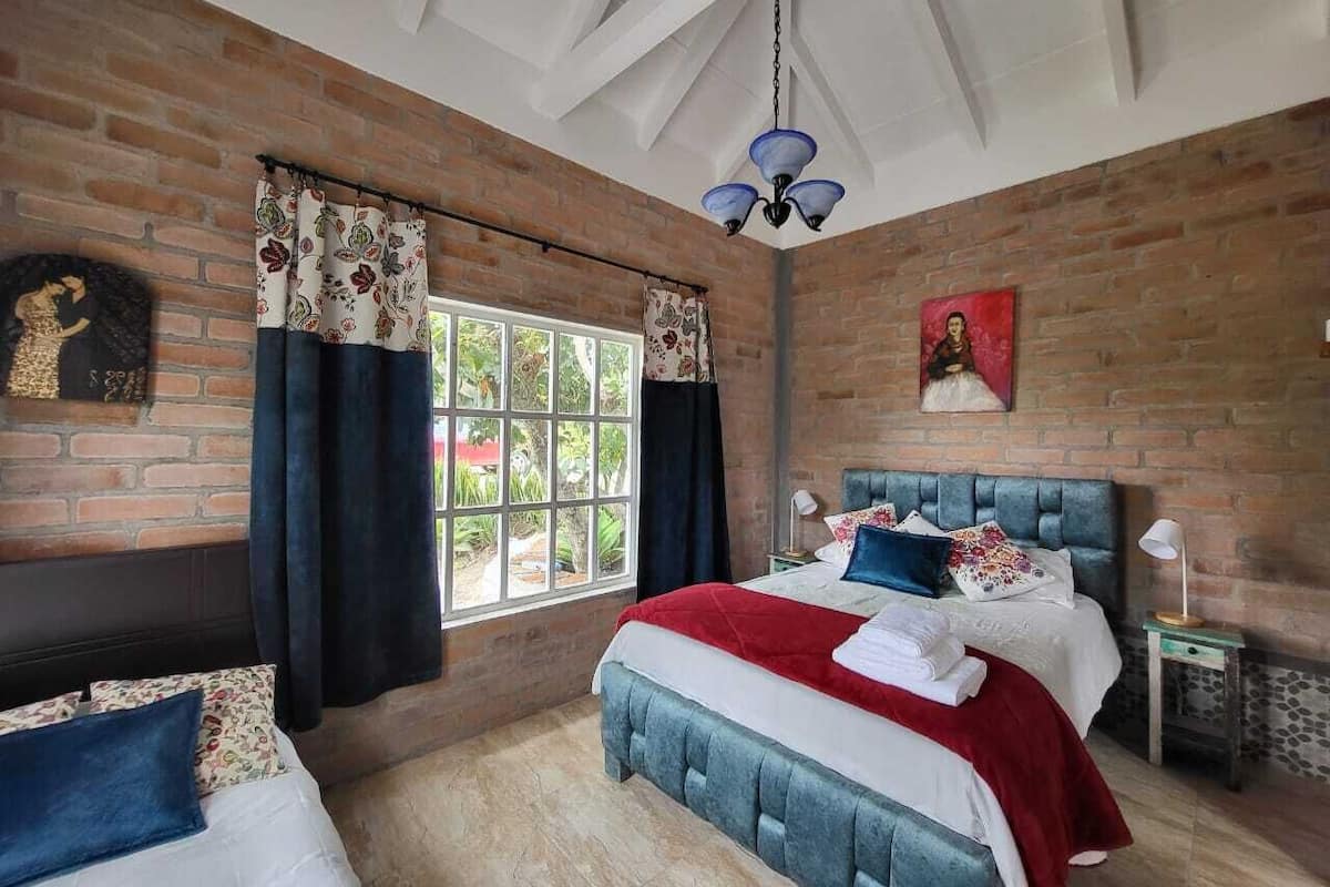 A bedroom with a brick wall, a large window, a bed with a red blanket, and a ceiling fan.