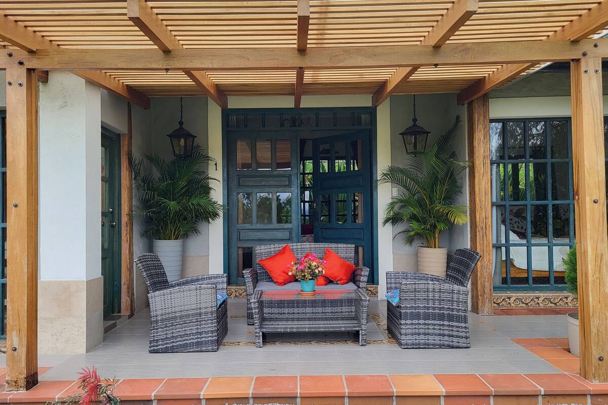 A covered outdoor seating area with wicker furniture, a central table with a vase of flowers, and a blue door with a decorative design.