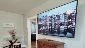 A room with a wooden dresser, a TV displaying a cityscape, a dining table with chairs, and a patterned rug. - Balcony Vibes and Blue Skies (Montgomery)